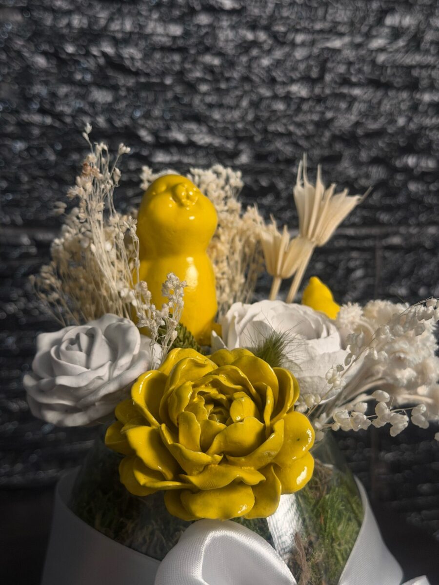Flor decorativa amarela em primeiro plano, com coelho amarelo, flores brancas e ramos claros ao fundo.