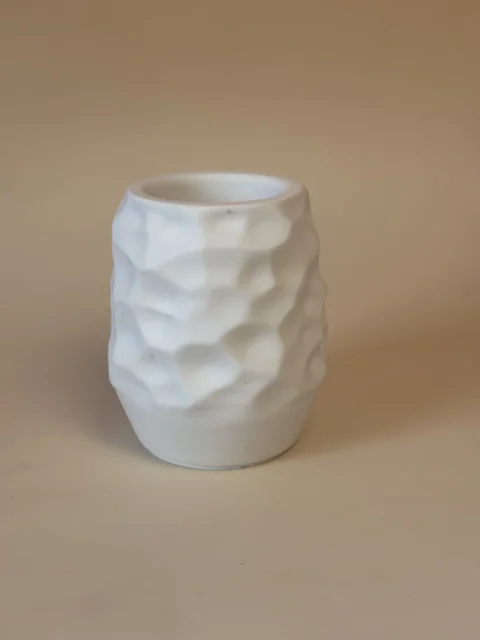 Jarra branca com abertura circular e superfície de facetas geométricas, sobre fundo bege.