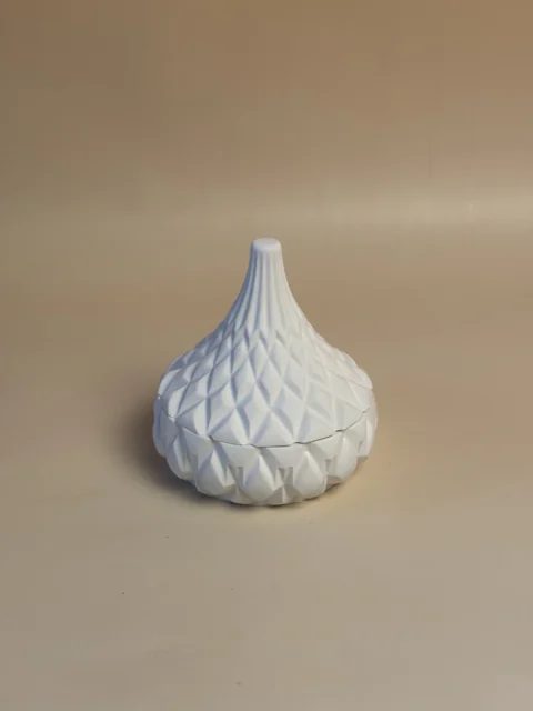 Pote branco em forma de pera com tampa pontiaguda e relevo geométrico, visto de um ângulo elevado.