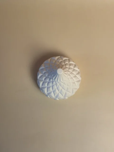 Pote branco em forma de pera, visto de cima, com relevo geométrico em torno da ponta central.