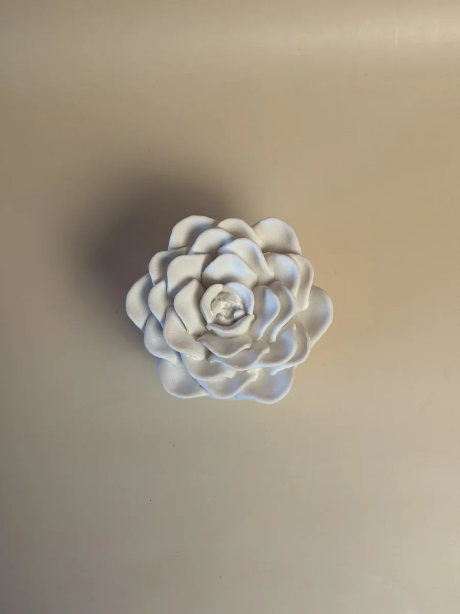 Pote branco com tampa em forma de rosa, visto de cima, mostrando as camadas de pétalas em relevo.