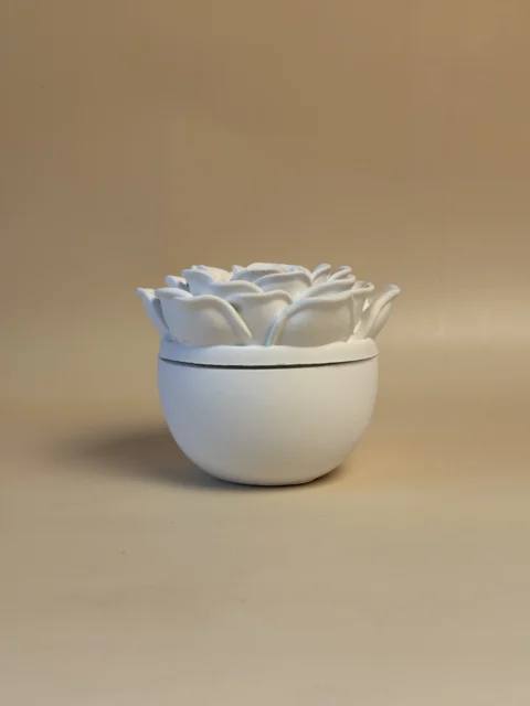 Vista frontal de um pote branco com base arredondada lisa e tampa decorada com uma rosa em relevo.