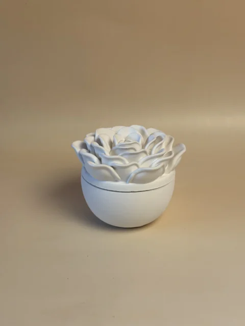 Pote branco arredondado com tampa em forma de rosa, visto de um ângulo elevado que destaca as pétalas.