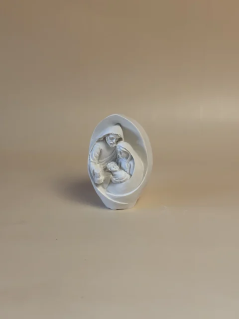 Pequena escultura branca da Sagrada Família, envolvida por uma forma oval semelhante a uma gruta.