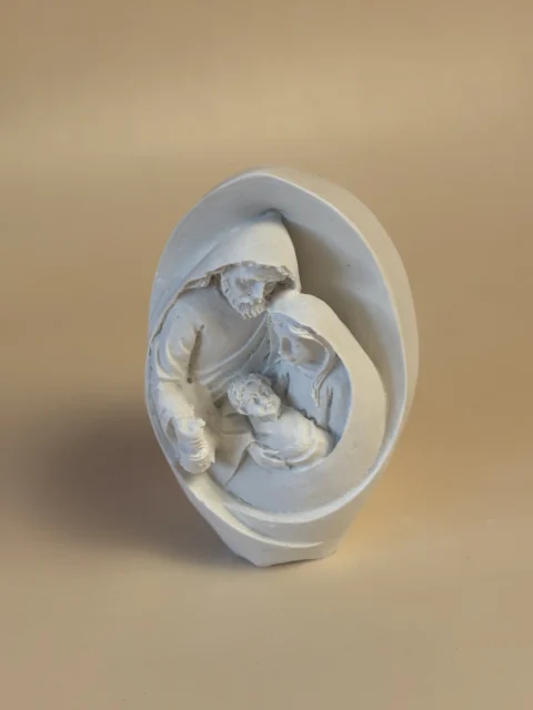 Pormenor da Sagrada Família numa gruta branca oval, com José e Maria inclinados sobre o Menino Jesus.