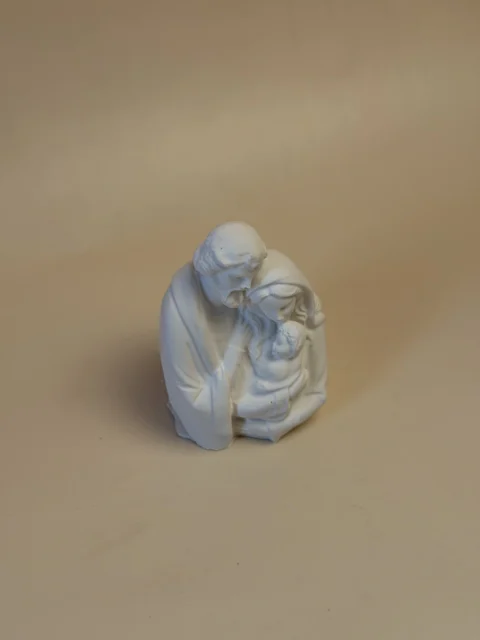 Escultura branca da Sagrada Família, vista de um ângulo elevado, com José inclinado sobre Maria e o Menino.