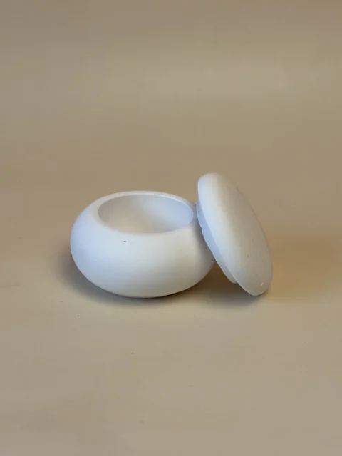 Pote branco arredondado com abertura circular e tampa lisa inclinada ao lado, visto de perto.